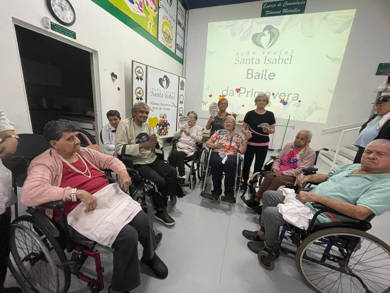 Idosos em cadeiras de rodas reunidos no salão decorado para o Baile de Primavera