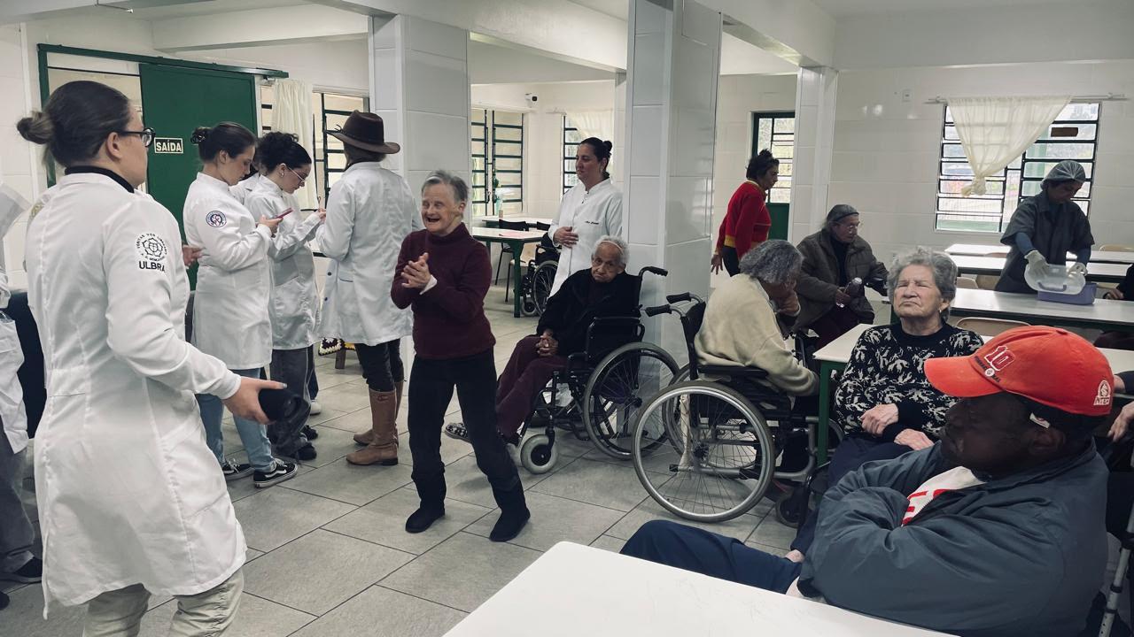 Estagiários de jaleco branco conversam com idosos no refeitório, alguns em cadeiras de rodas