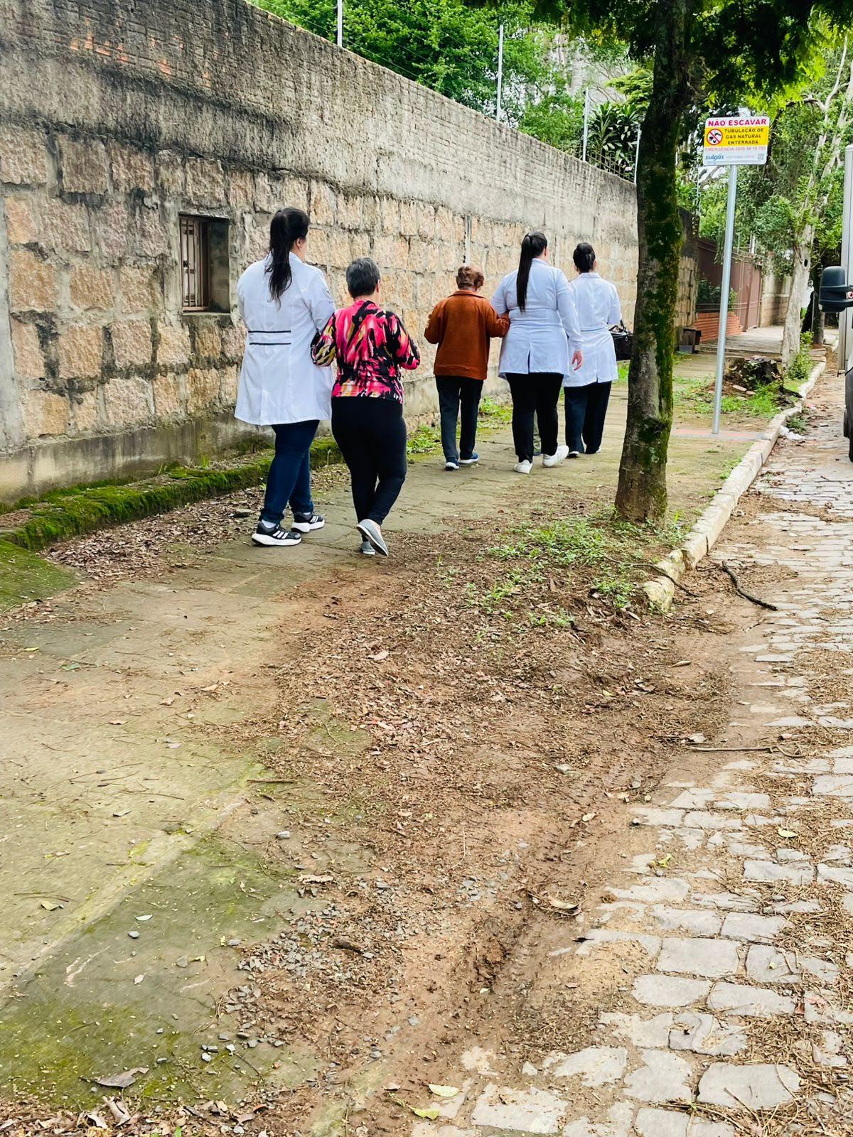Estagiárias caminham com idosas por uma rua arborizada, ao lado de um muro de pedra