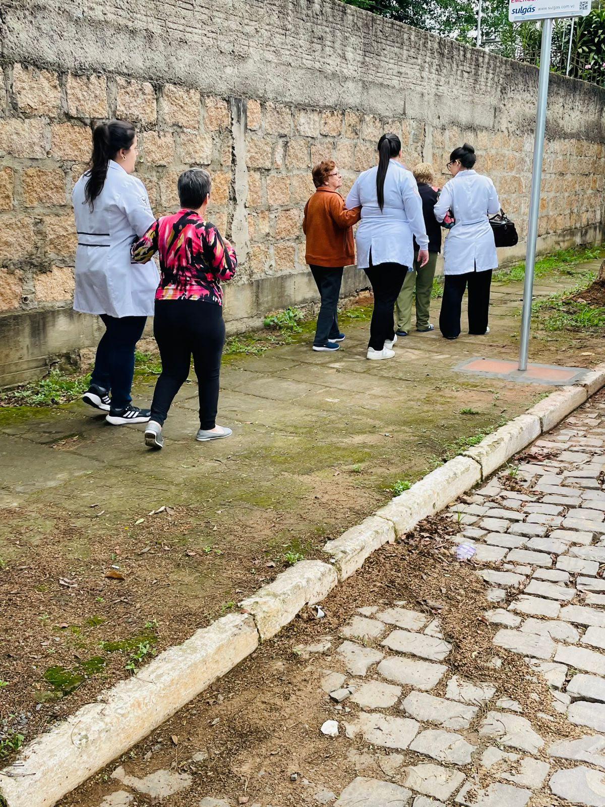 Grupo de estagiárias e idosas caminha pela calçada ao lado de um muro de pedra