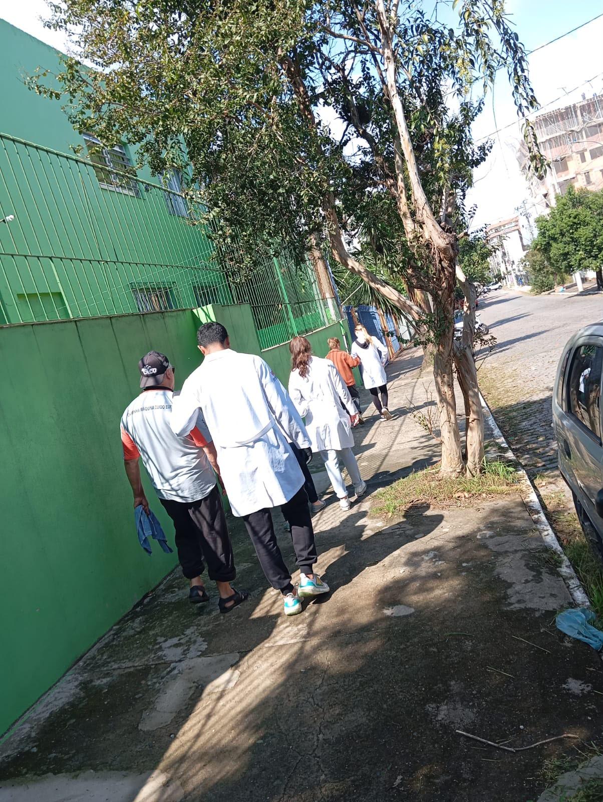 Estagiários de jaleco acompanham idosos em caminhada pela calçada