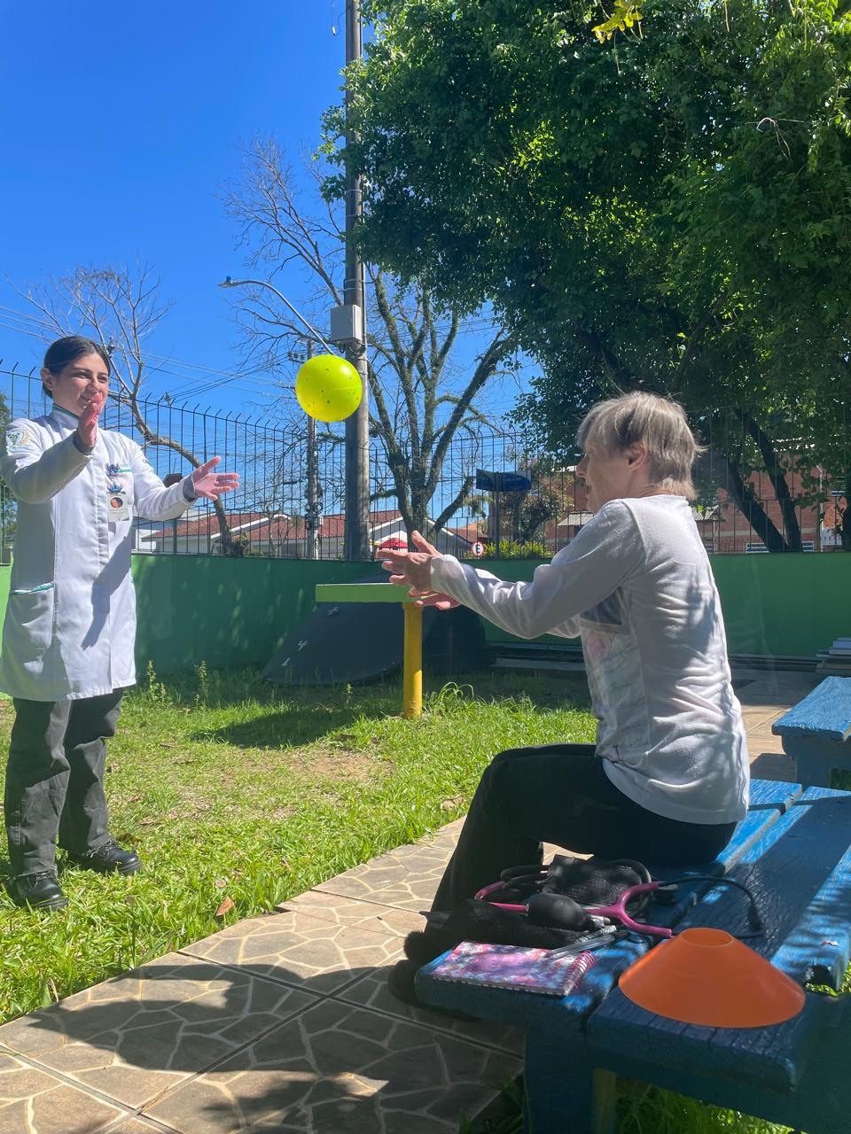 Estagiária joga uma bola para uma idosa sentada em um banco do jardim
